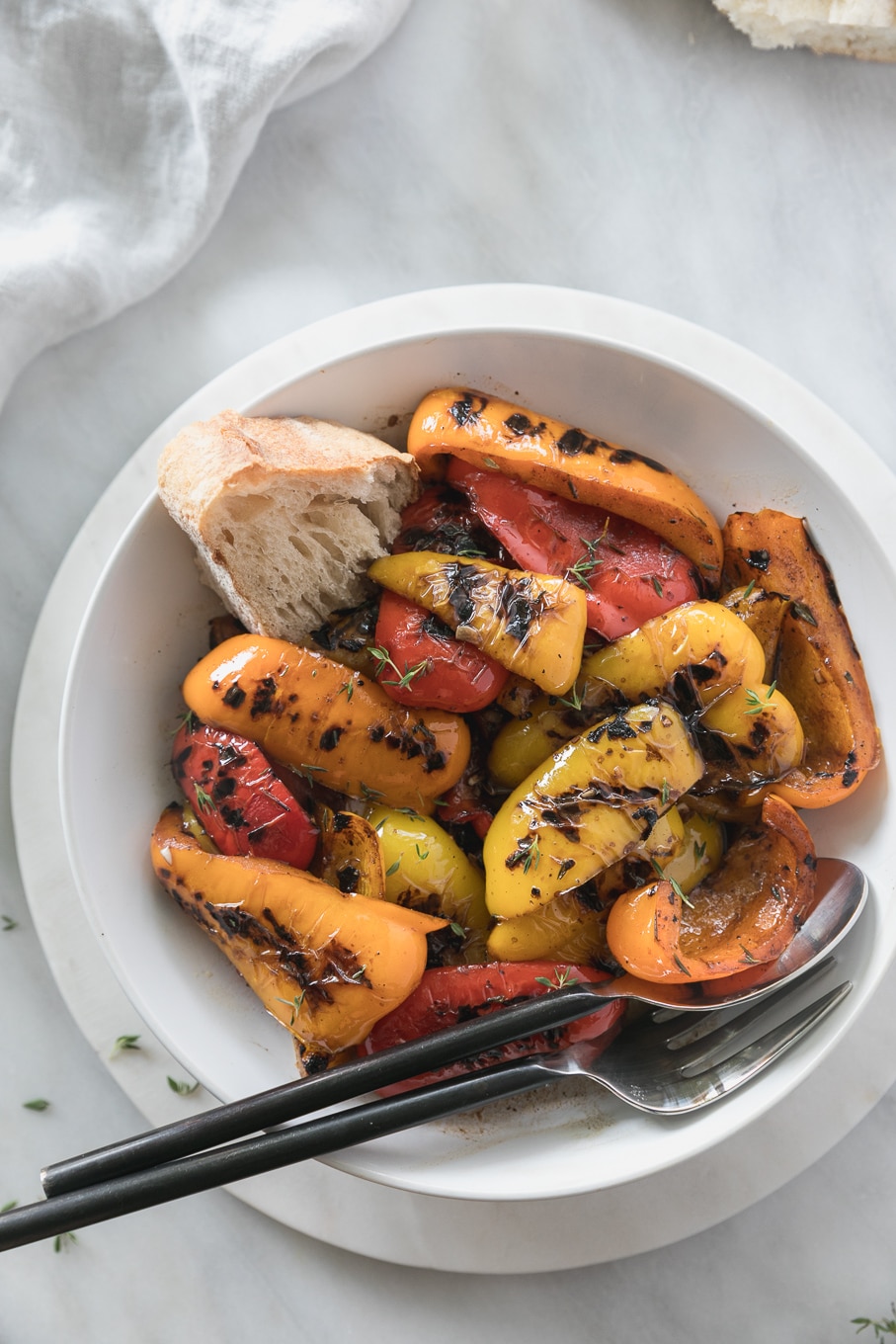 Overhead shot of a bowl of grilled peppers with a large spoon and fork in the bowl, as well as a half piece of bread